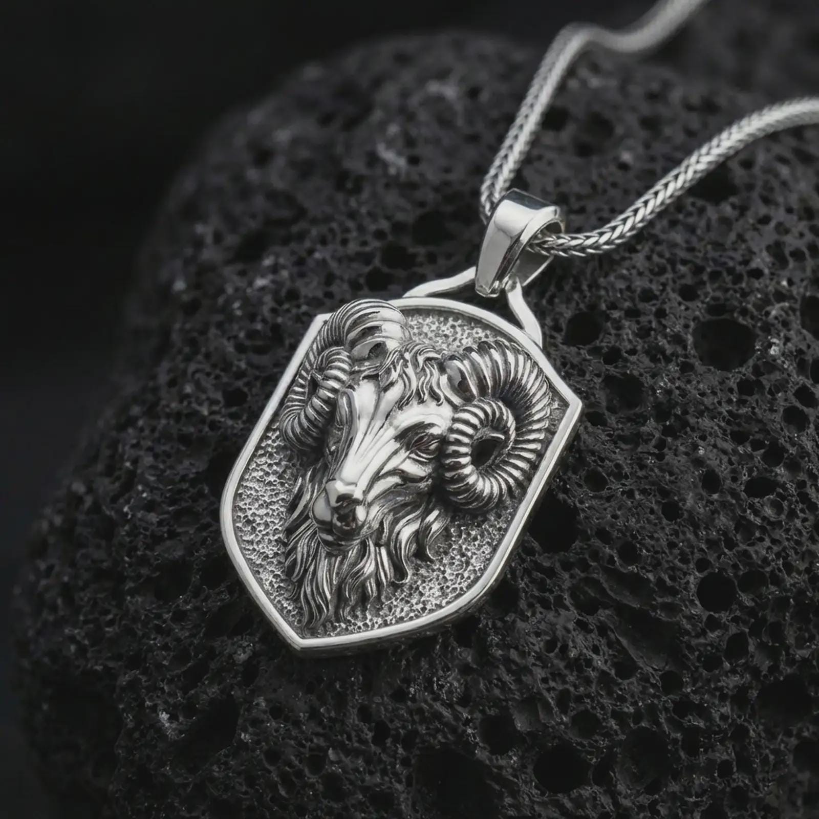 Polished silver ram head shield pendant necklace displayed on black volcanic rock, shown in a refined macro product shot with curled horns, sculpted Aries face detail, and angular shield frame.
