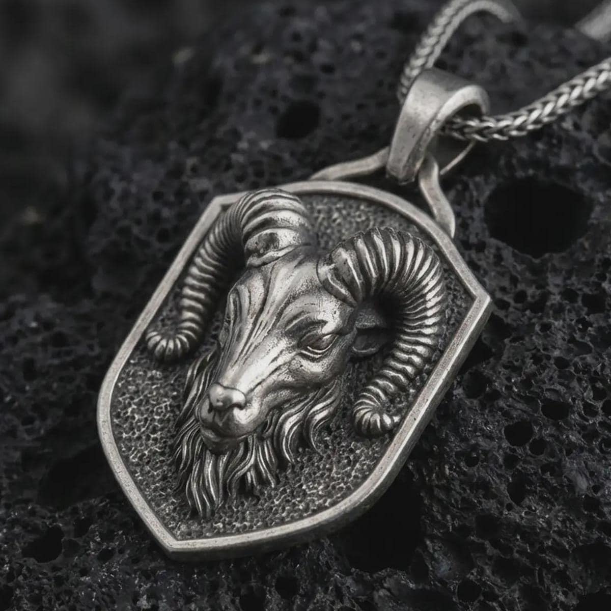Ram head shield pendant necklace in oxidized sterling silver resting on black volcanic stone, shown in a detailed macro product photograph with curved horns and a textured shield frame.