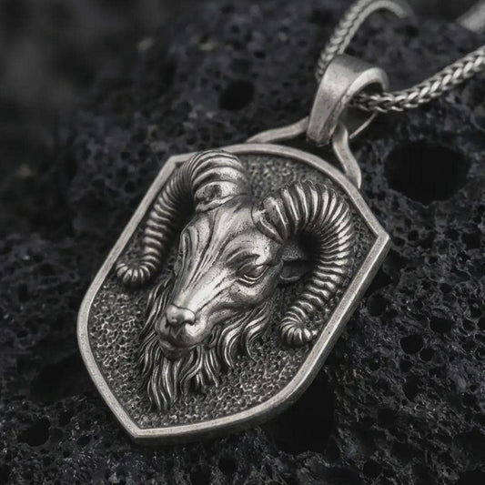Ram head shield pendant necklace in oxidized sterling silver resting on black volcanic stone, shown in a detailed macro product photograph with curved horns and a textured shield frame.