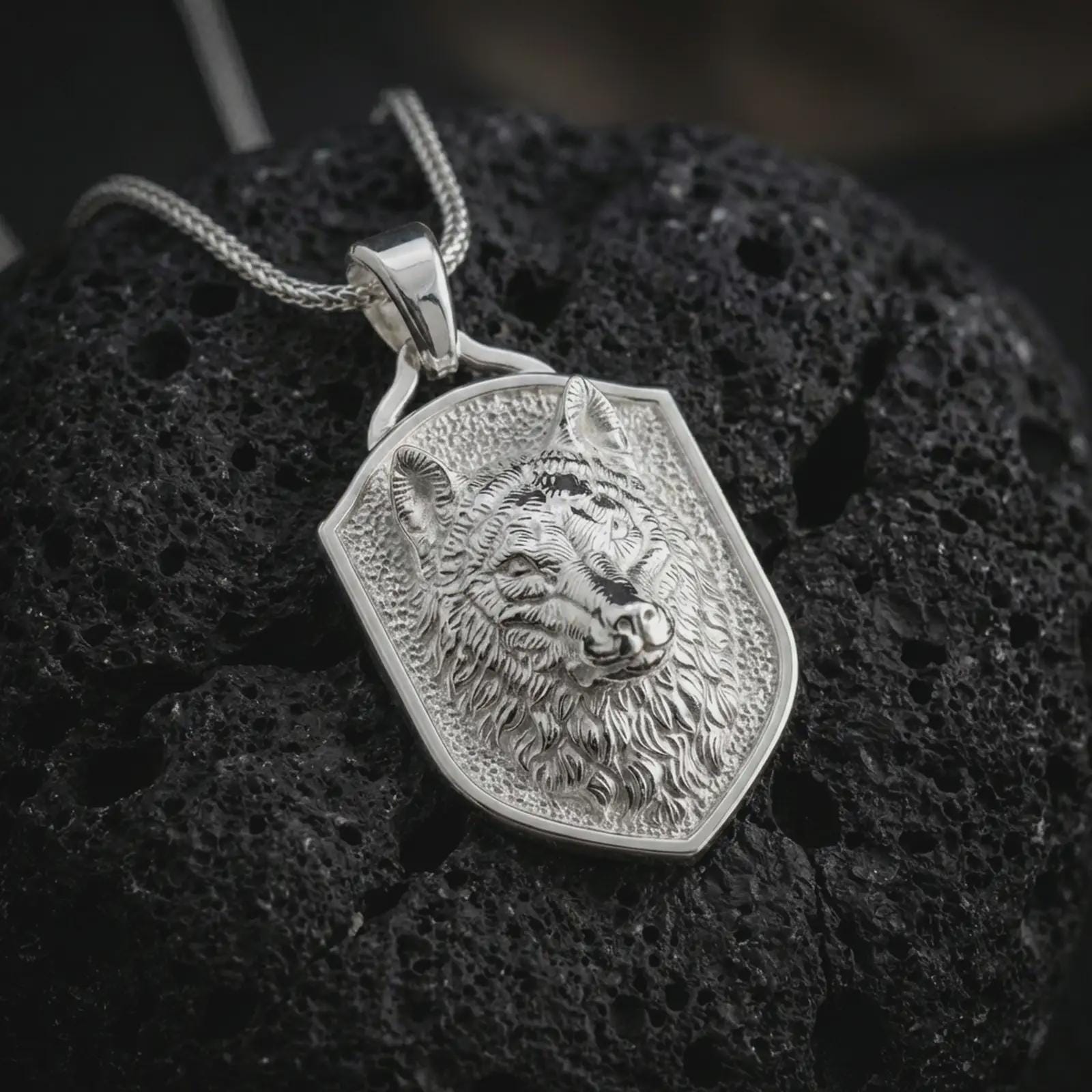 Wolf shield pendant necklace in polished silver displayed on dark volcanic rock, captured in a close-up studio image that highlights the sculpted wolf face and bold protective shield silhouette.