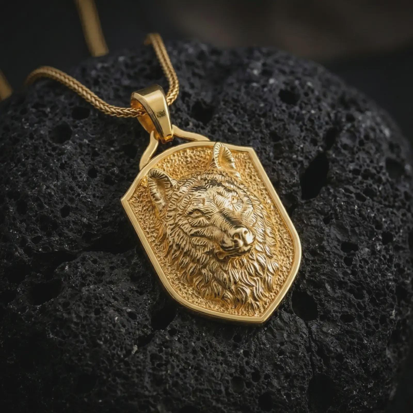 Wolf shield pendant necklace with a gold plated finish placed on porous volcanic stone, photographed in a refined macro composition with strong predator detail and symbolic warrior styling.