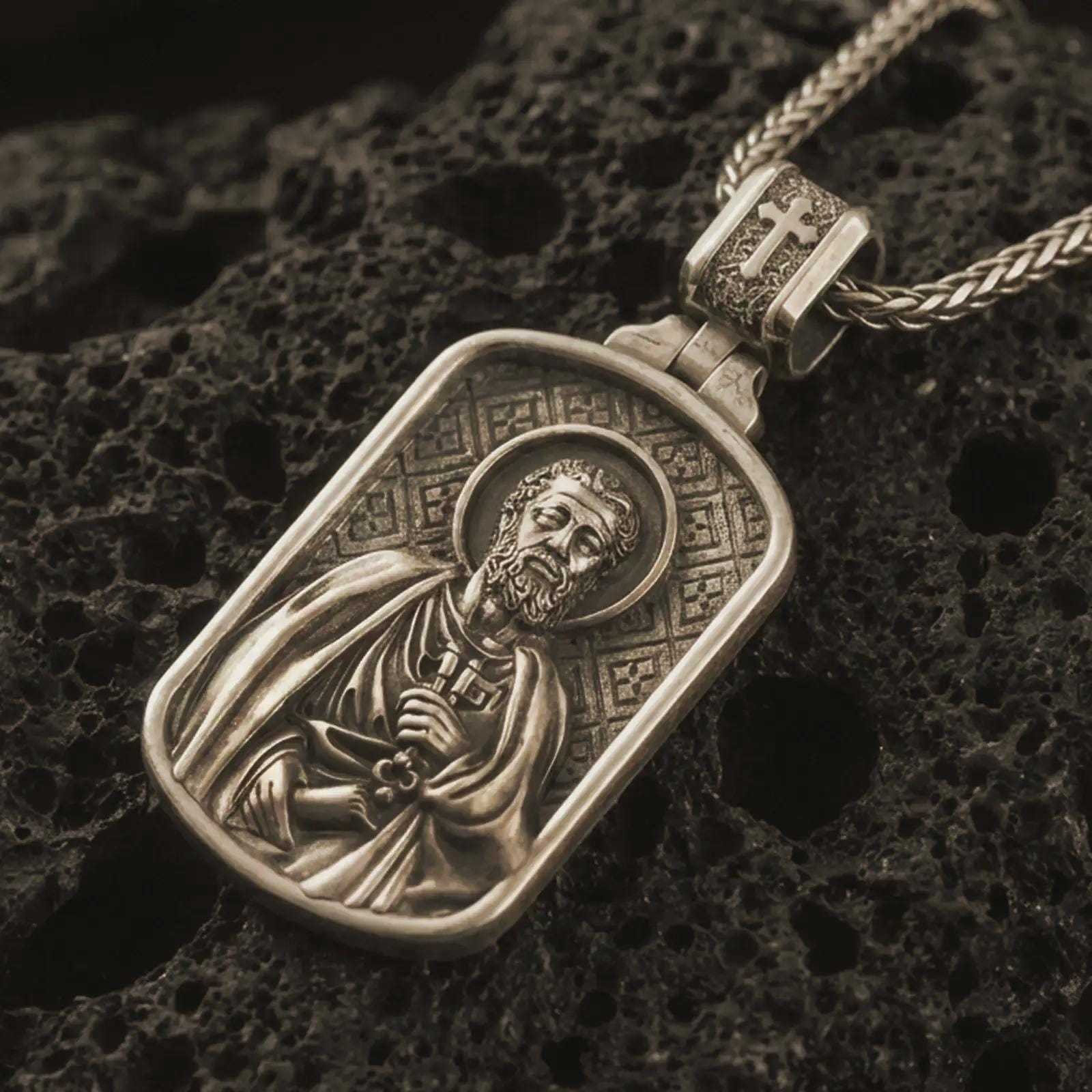 Bronze Saint Peter dog tag pendant necklace with an oxidized finish resting on textured volcanic rock, captured in a close-up premium studio composition.