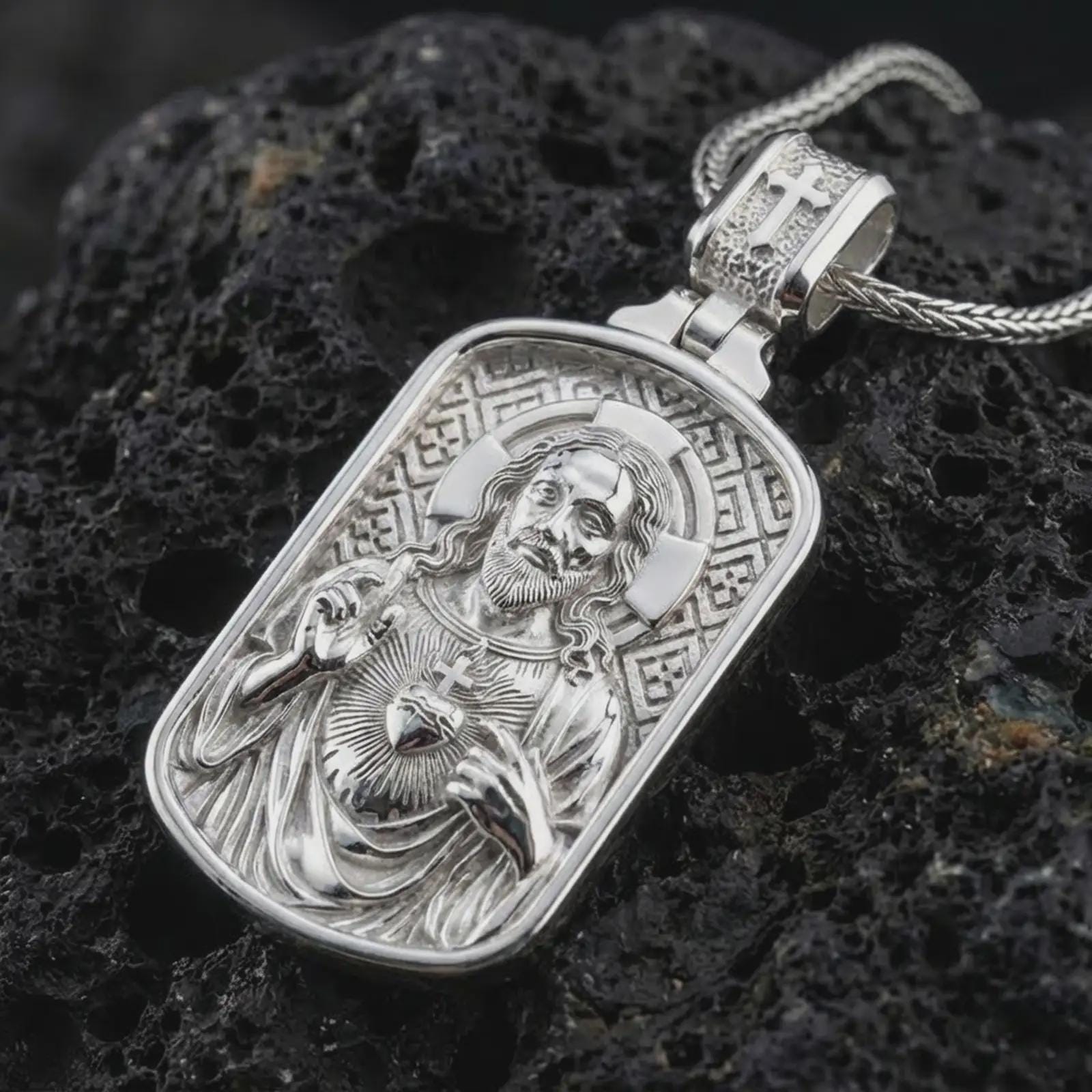 Polished silver Sacred Heart pendant necklace displayed on black volcanic rock, shown in a refined macro product shot with detailed Christ figure and engraved background pattern.