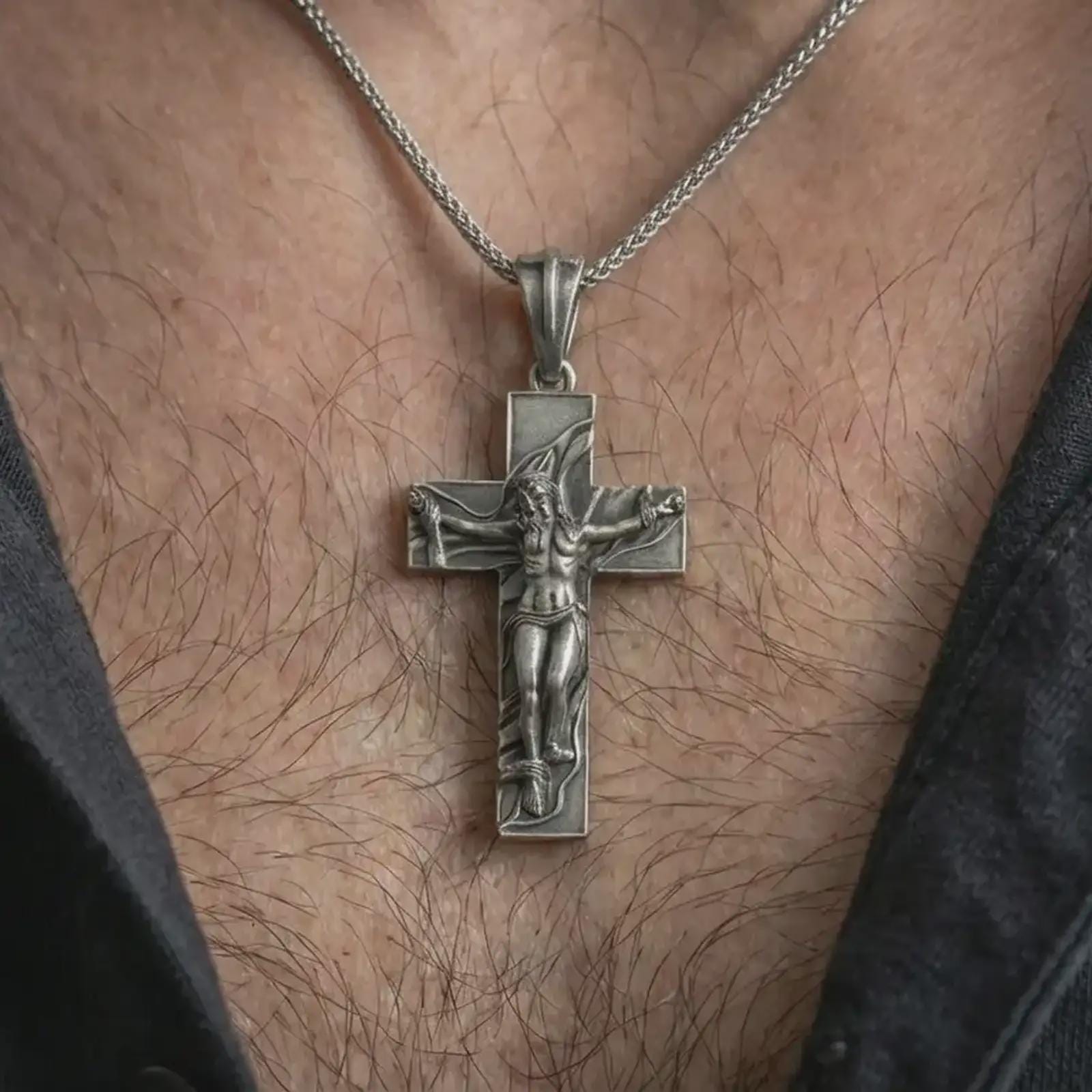 Oxidized symbolic cross pendant worn on the neck, styled in a dark lifestyle portrait with an open shirt and masculine presentation.