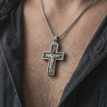 Load image into Gallery viewer, Oxidized Cristo Protetor cross pendant necklace worn on the neck, styled in a dark lifestyle portrait with an open shirt and masculine presentation.

