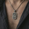 Load image into Gallery viewer, Oxidized Saint Paul dog tag pendant necklace worn on the neck, styled in a dark lifestyle portrait with an open shirt and masculine presentation.
