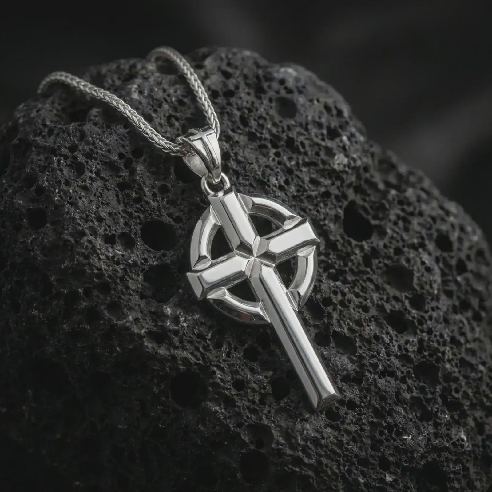 Polished silver Celtic cross pendant necklace resting on textured volcanic rock, shown in a premium macro product shot emphasizing its clean Christian silhouette and solid geometric design.