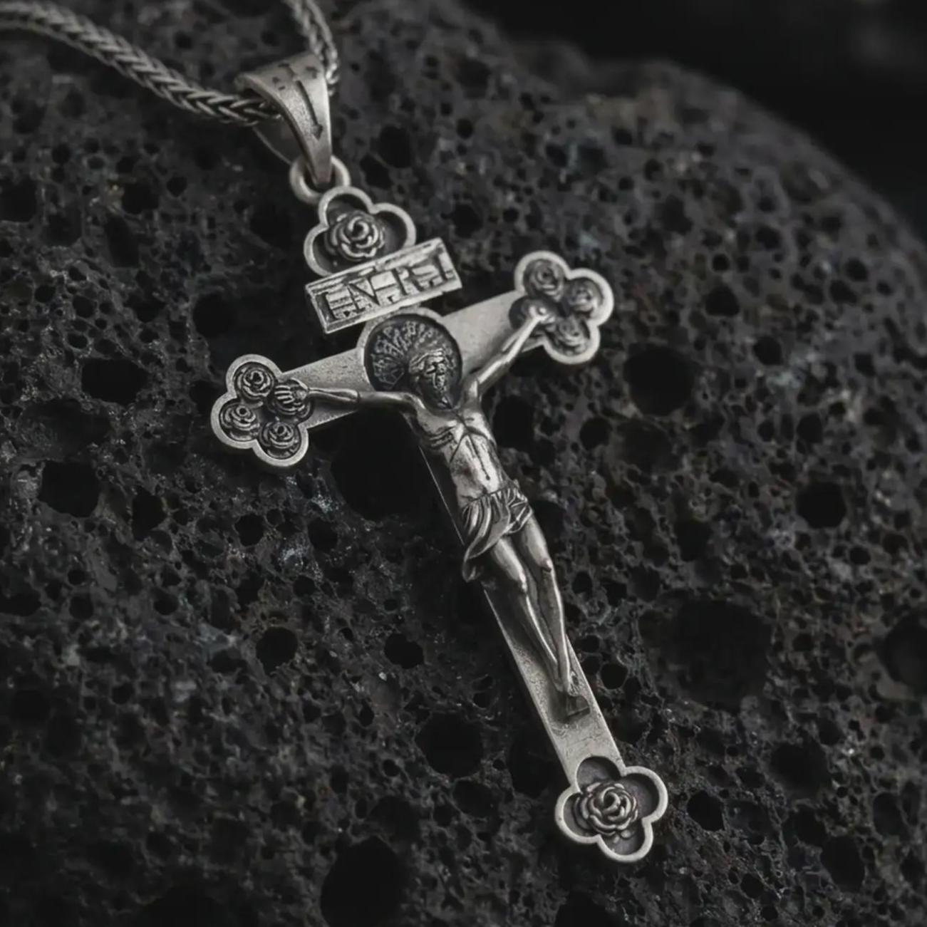 Sterling silver crucifix pendant necklace with an oxidized finish displayed on black volcanic stone, captured in a close-up studio image highlighting the detailed Jesus figure and floral cross tips.
