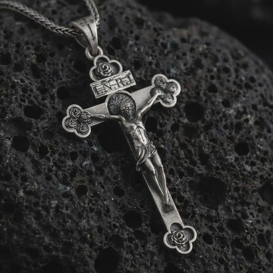 Sterling silver crucifix pendant necklace with an oxidized finish displayed on black volcanic stone, captured in a close-up studio image highlighting the detailed Jesus figure and floral cross tips.