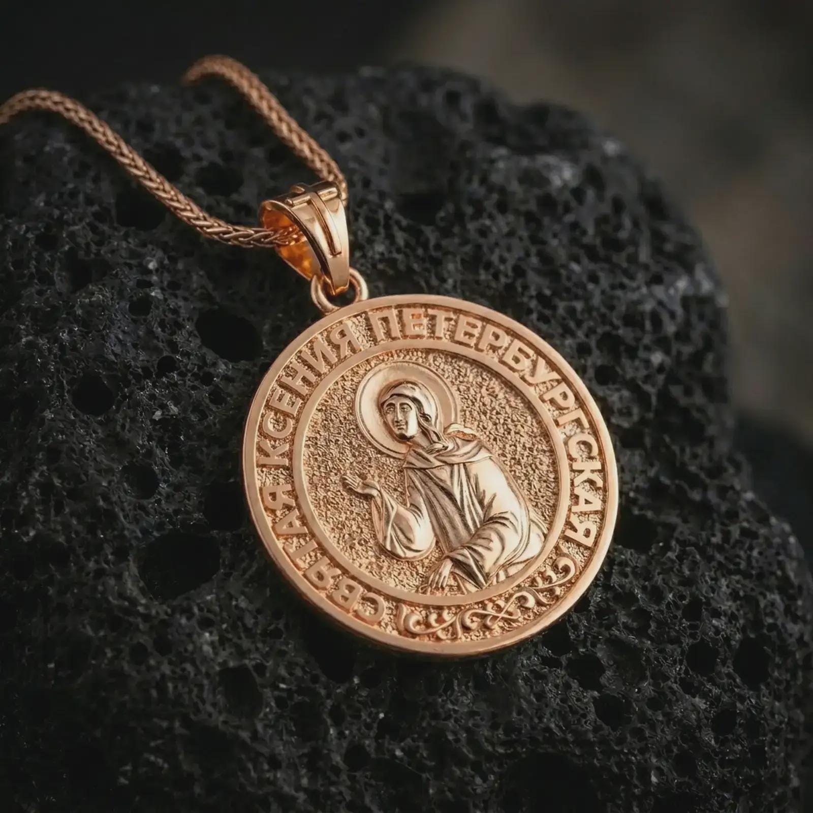 Rose gold plated Saint Xenia pendant necklace photographed on porous volcanic stone, showcasing the Saint Ksenia icon, ornamental border, and refined spiritual craftsmanship.