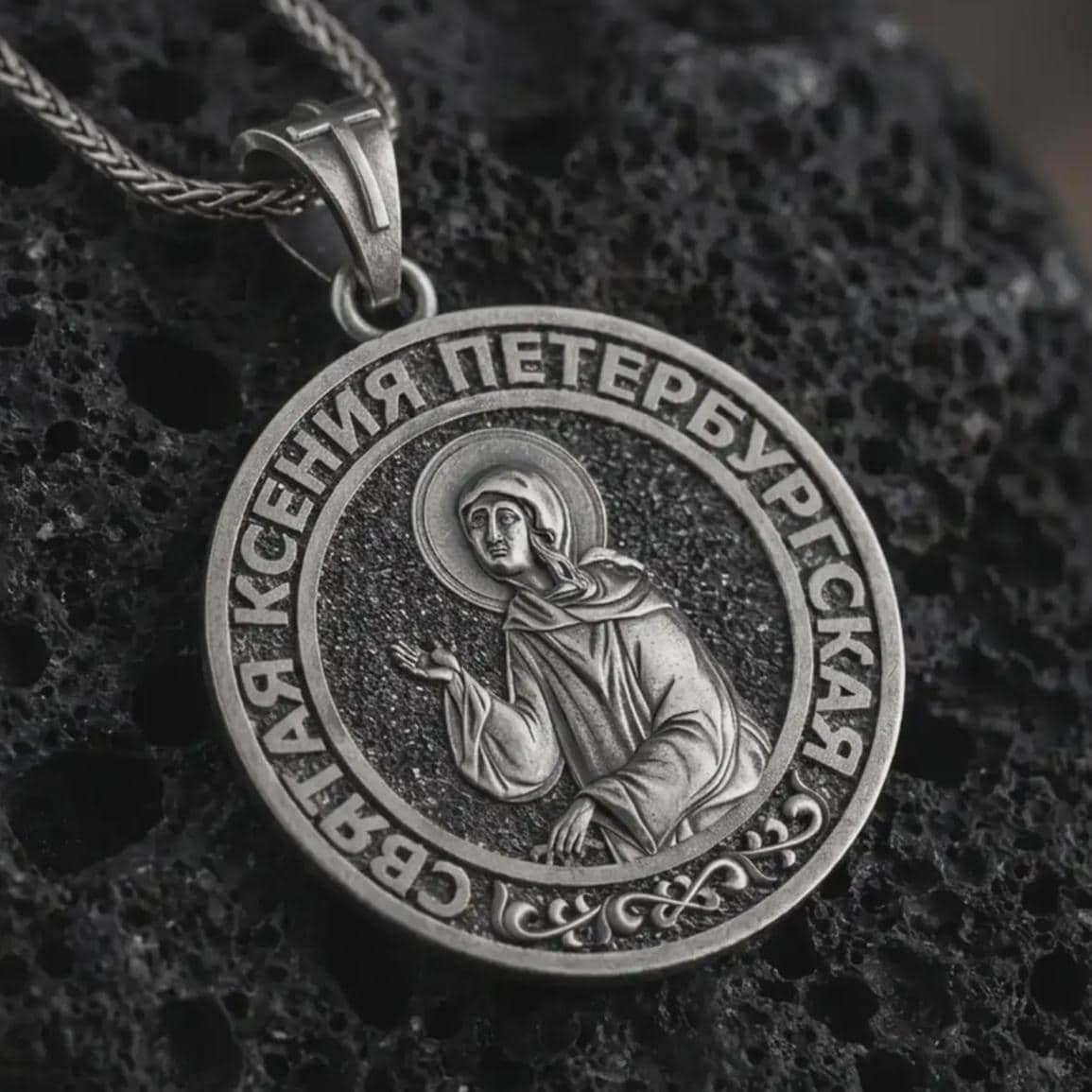 Oxidized Saint Xenia pendant necklace displayed on black volcanic stone, featuring a round Orthodox medal with Saint Ksenia of Petersburg and Cyrillic inscription in a detailed macro product shot.
