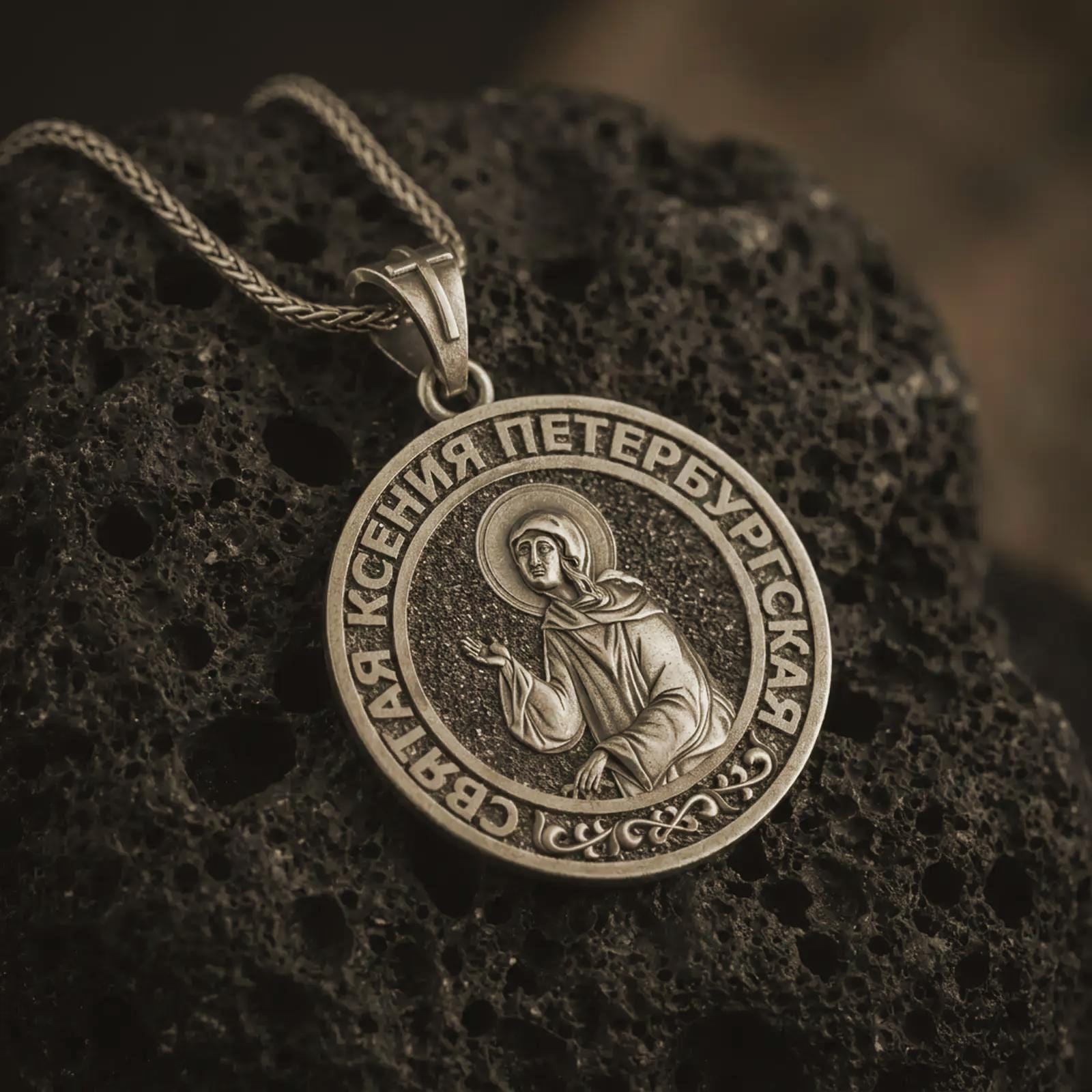 Bronze Saint Xenia pendant necklace displayed on black volcanic stone, featuring a round Orthodox medal with Saint Ksenia of Petersburg and Cyrillic inscription in a detailed macro product shot.