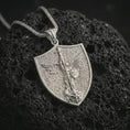 Load image into Gallery viewer, Polished silver Saint Michael pendant necklace resting on dark volcanic rock, captured in a premium close-up that highlights the angel wings, spear, and protective shield motif.

