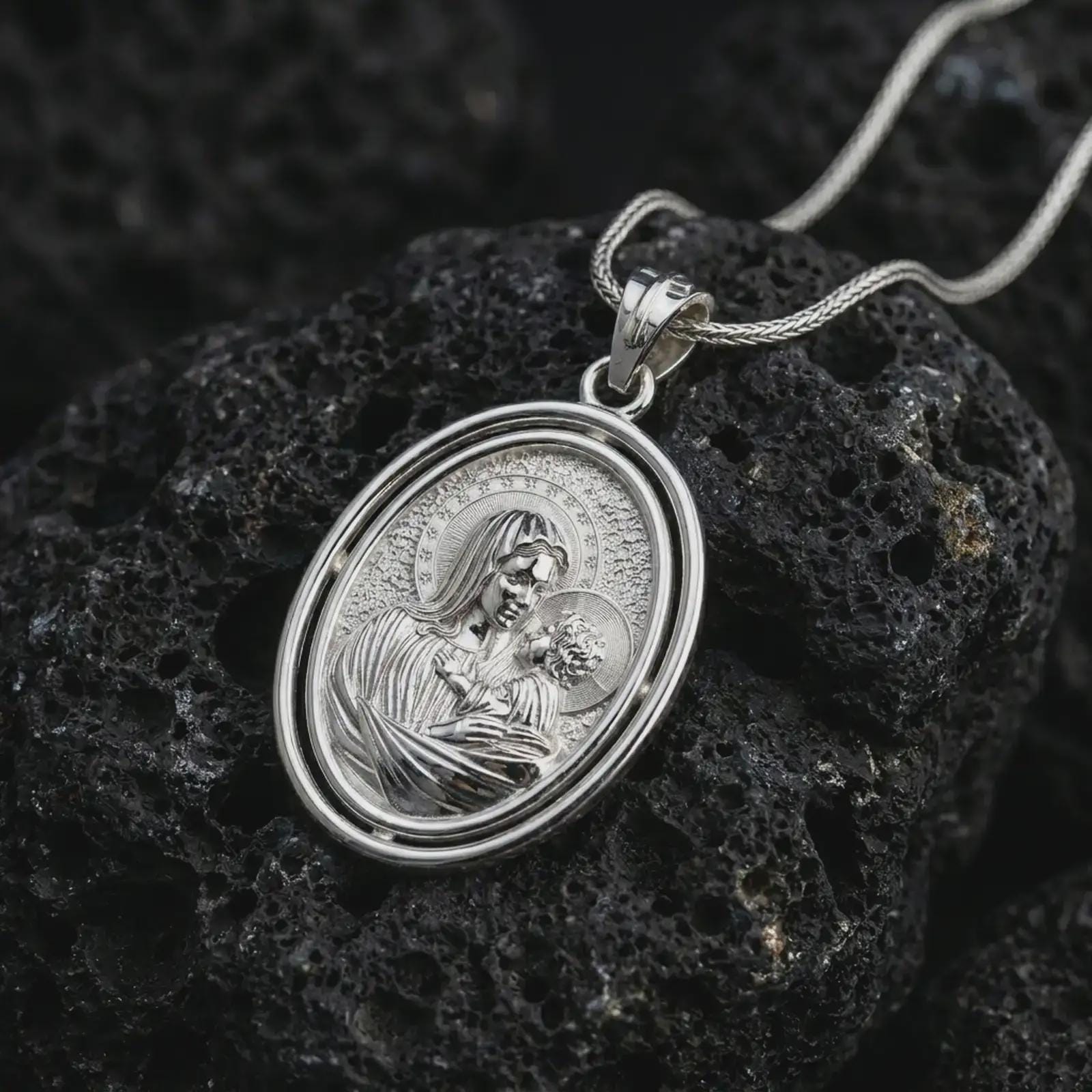 Polished silver Madonna and Child pendant necklace displayed on dark volcanic rock, highlighting the sacred mother and infant motif within an elegant oval religious medal design.