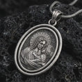 Load image into Gallery viewer, Oxidized Madonna and Child pendant necklace resting on black volcanic stone, featuring an oval double-frame medallion with the Virgin Mary holding baby Jesus in a detailed devotional relief.
