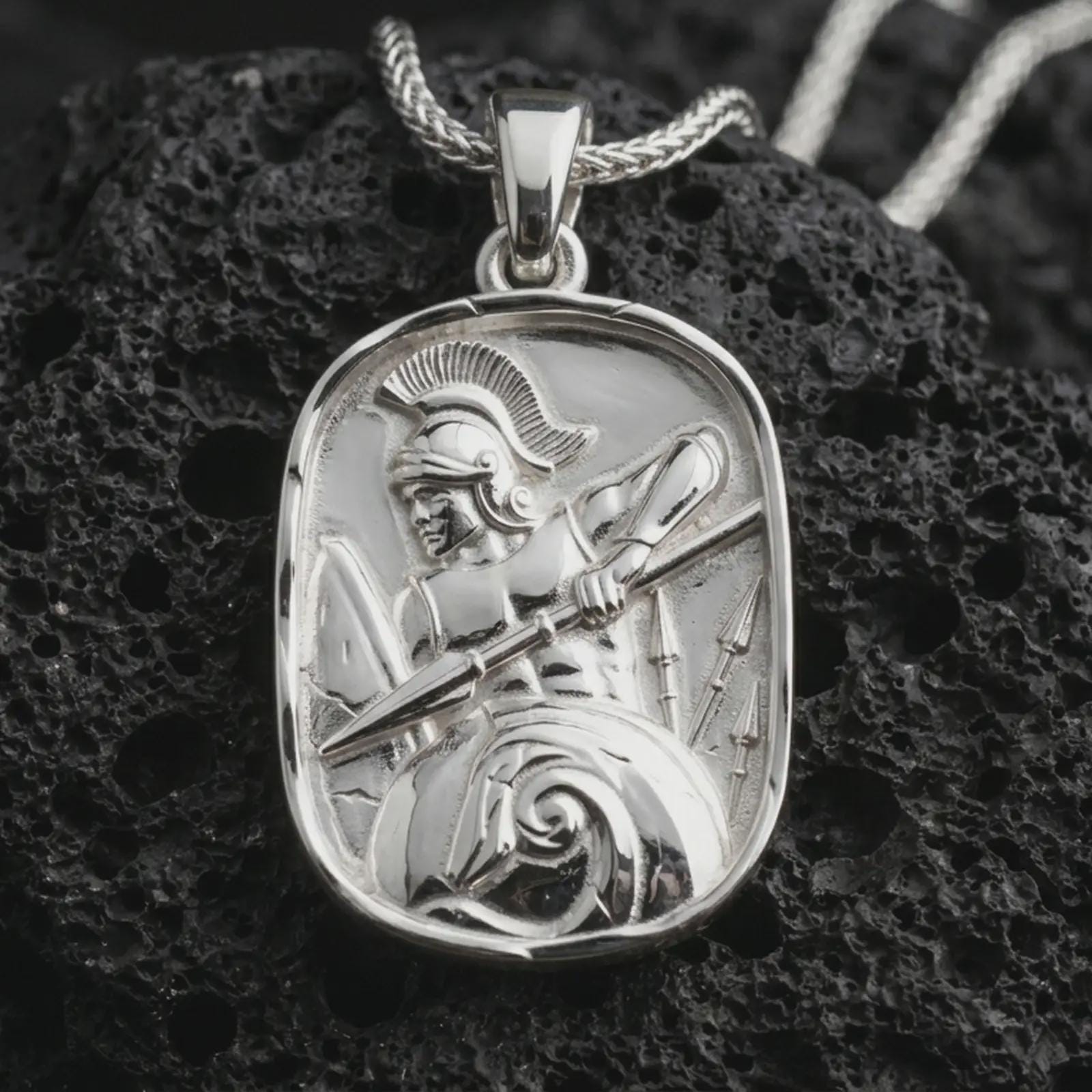 Polished silver Spartan warrior pendant necklace displayed on black volcanic rock, shown in a refined macro product shot with detailed helmet, spear, and shield relief.