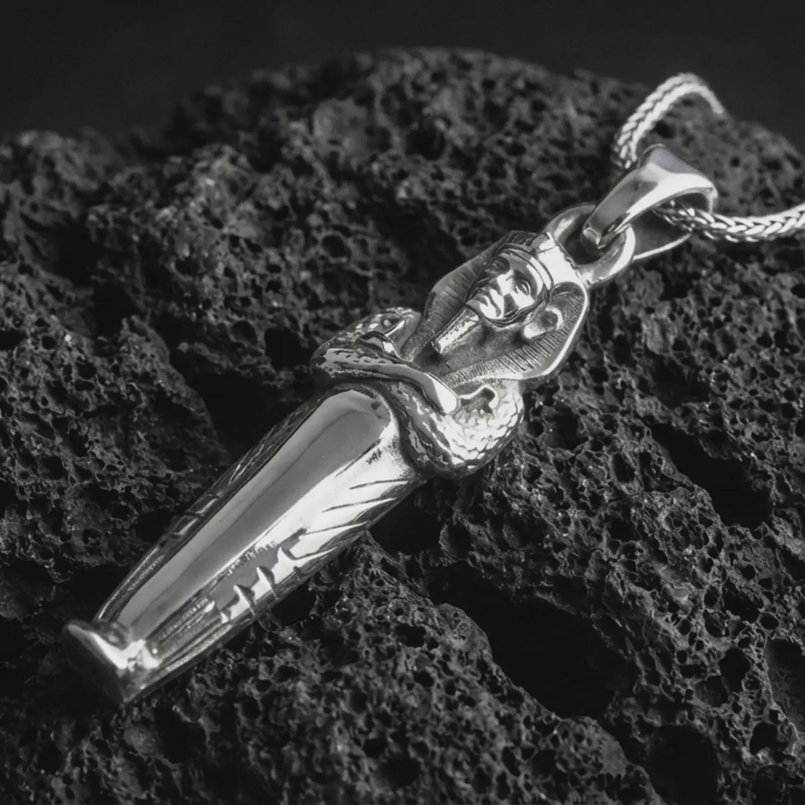 Polished silver King Tut pendant necklace displayed on black volcanic rock, shown in a refined macro product shot with detailed pharaoh head and serpent relief.