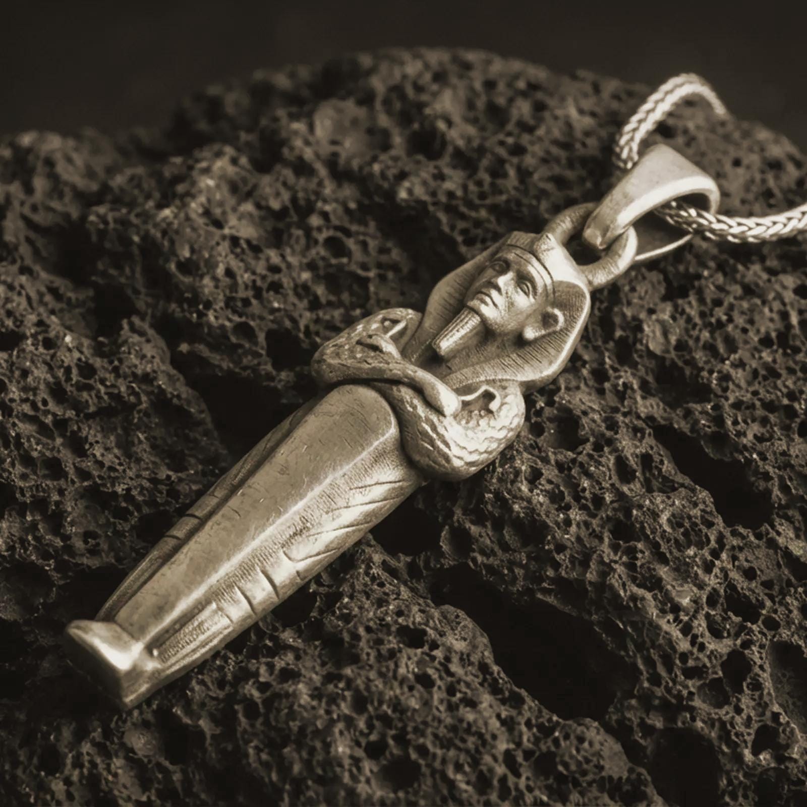 Bronze King Tut pendant necklace with an oxidized finish resting on textured volcanic rock, captured in a close-up premium studio composition.