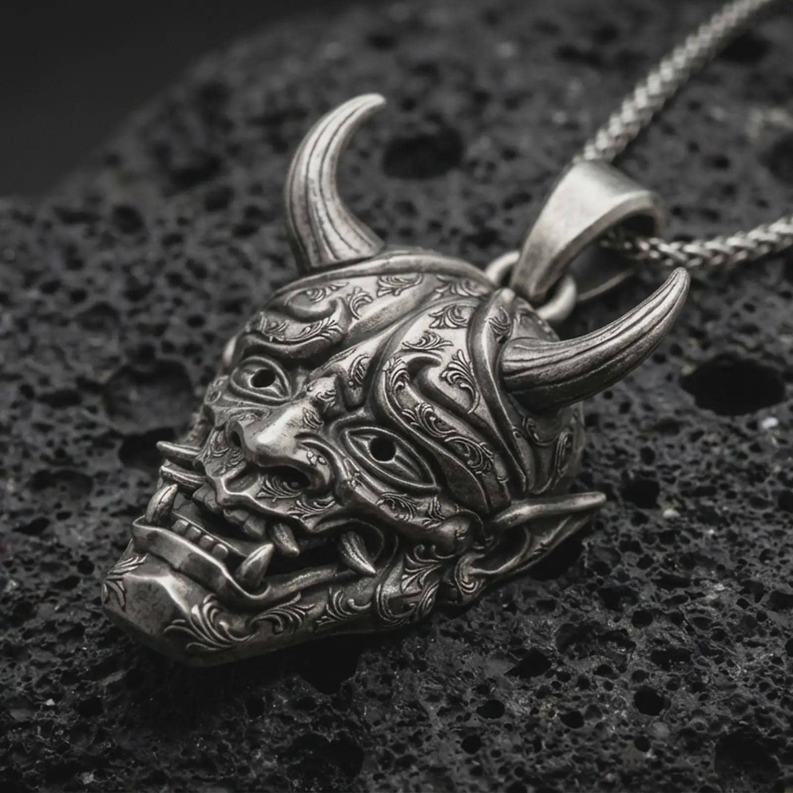Sterling silver Oni mask pendant necklace with an oxidized finish resting on textured volcanic rock, captured in a close-up premium studio composition.