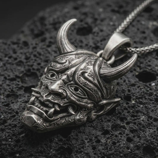 Sterling silver Oni mask pendant necklace with an oxidized finish resting on textured volcanic rock, captured in a close-up premium studio composition.