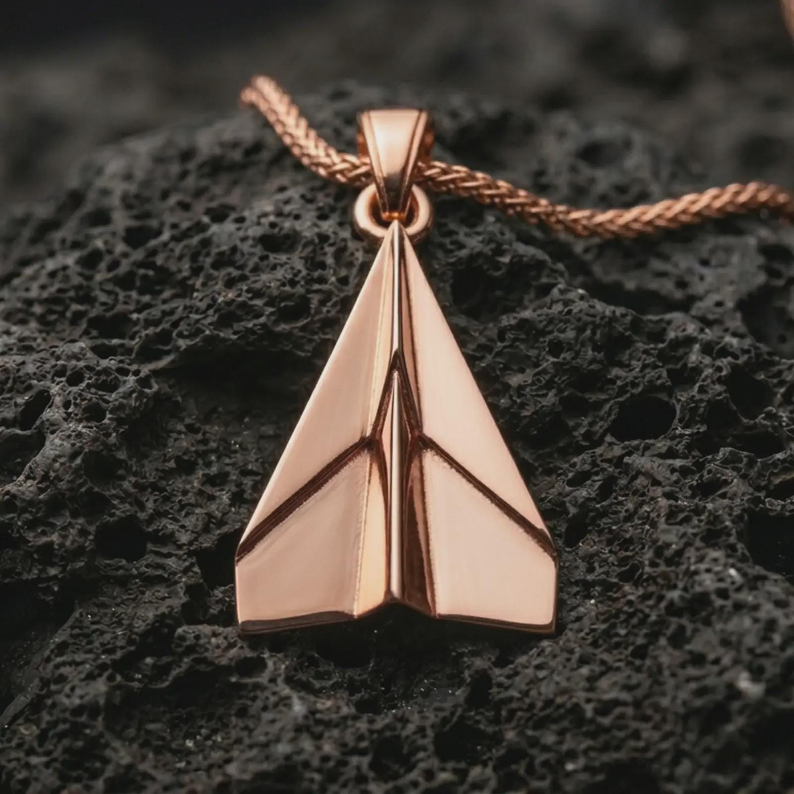 Rose gold plated origami airplane pendant necklace on dark volcanic rock, photographed in a detailed premium studio macro view.