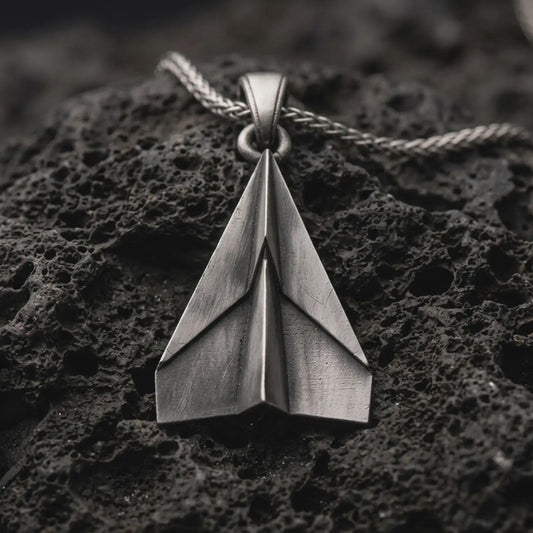 Sterling silver origami airplane pendant necklace with an oxidized finish resting on textured volcanic rock, captured in a close-up premium studio composition.