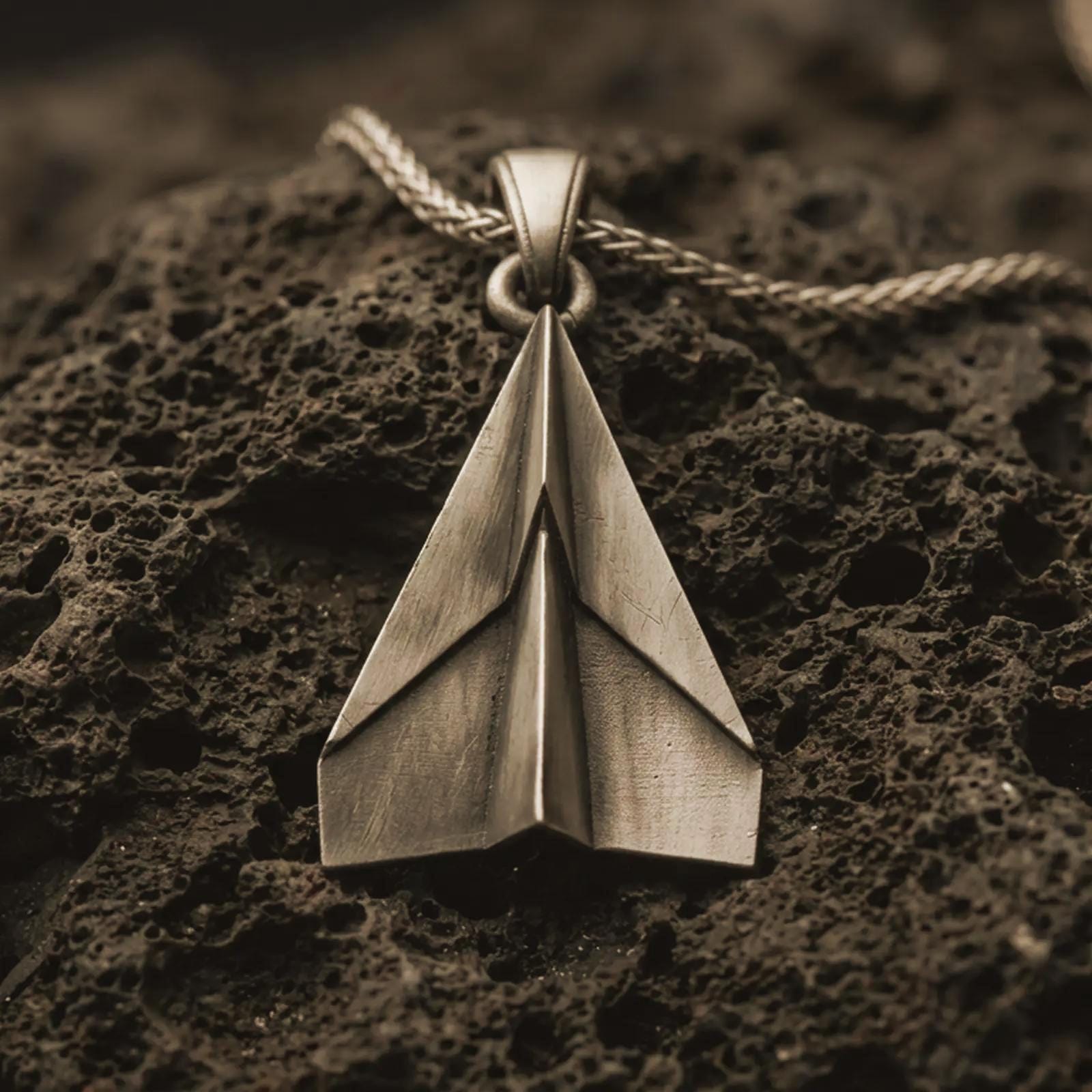 Bronze origami airplane pendant necklace with an oxidized finish resting on textured volcanic rock, captured in a close-up premium studio composition.