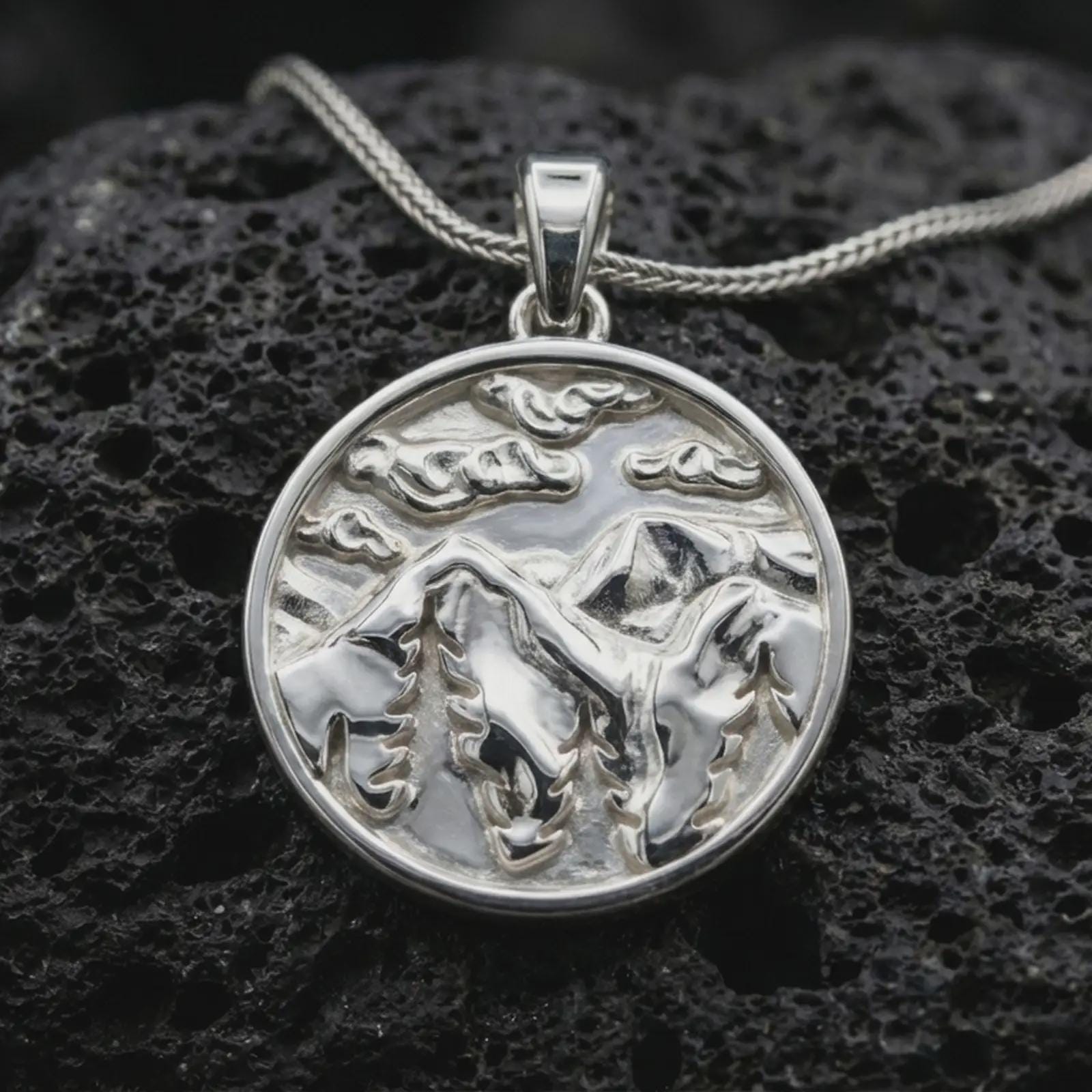Polished silver mountain landscape pendant necklace displayed on black volcanic rock, shown in a refined macro product shot with layered peaks, pine tree silhouettes, and cloud relief.