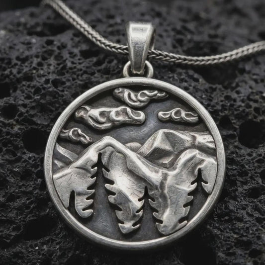 Sterling silver mountain landscape pendant necklace with an oxidized finish resting on textured volcanic rock, captured in a close-up premium studio composition.