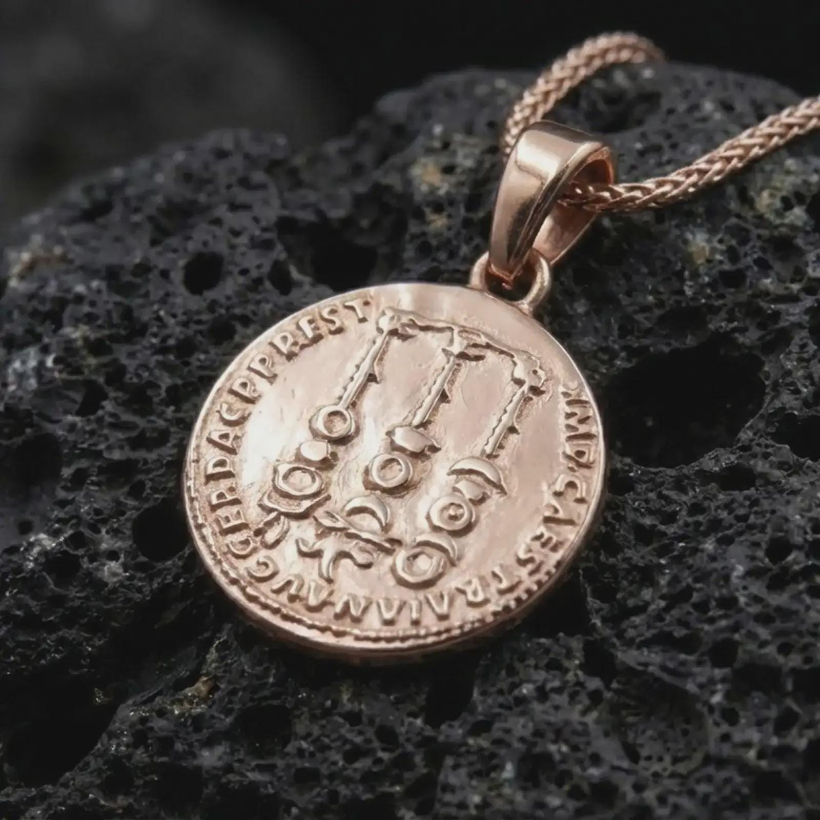 Rose gold plated Tiberius coin pendant necklace presented on textured volcanic stone, showcasing the round Roman coin silhouette and emperor bust in a refined macro studio shot.