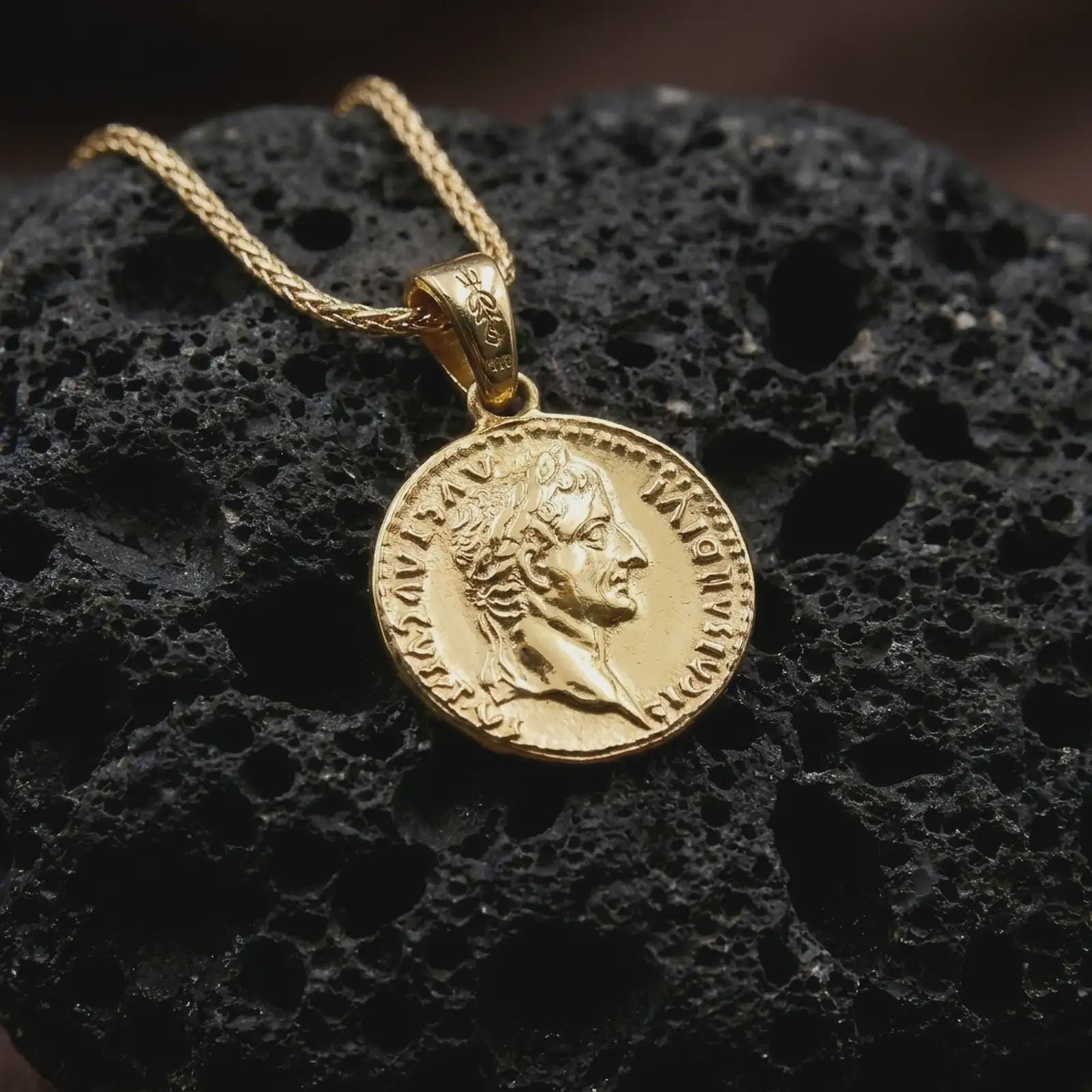 Gold plated Tiberius coin pendant necklace photographed on porous black volcanic stone, emphasizing the imperial side-profile relief and engraved ancient Roman lettering in a luxury jewelry composition.