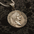 Load image into Gallery viewer, Bronze Tiberius coin pendant necklace resting on black volcanic stone, featuring a Roman emperor profile and antique coin-style inscription in a detailed macro product photograph.
