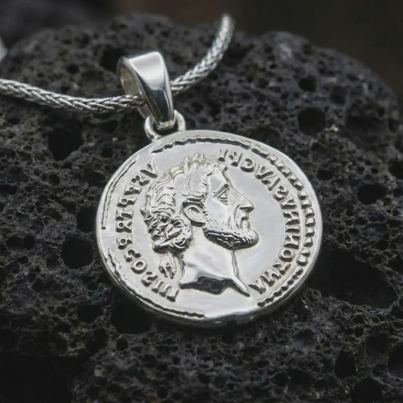 Polished silver Roman coin pendant necklace displayed on dark volcanic rock, shown in a premium close-up image that highlights the classical emperor portrait and worn ancient coin texture.