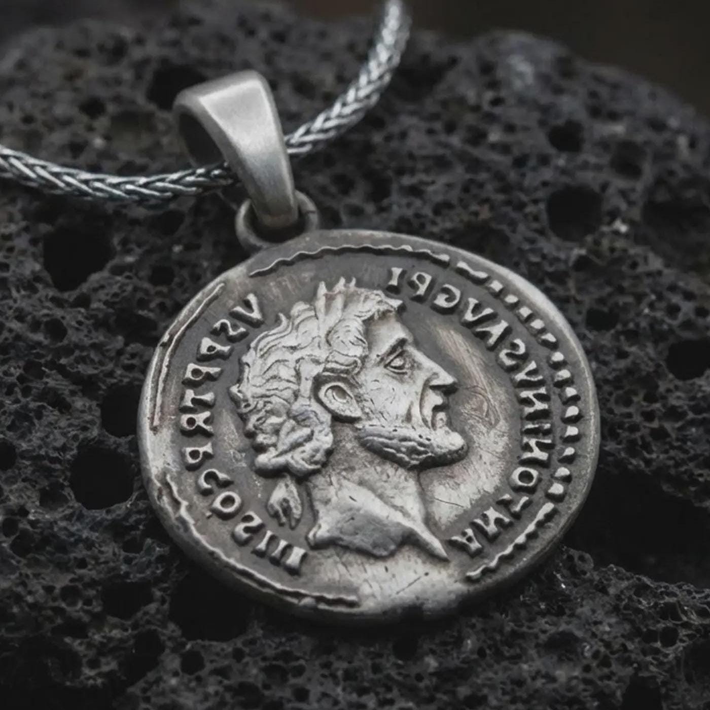 Oxidized Roman emperor coin pendant necklace resting on black volcanic stone, featuring a side profile imperial bust and antique Latin inscription in a detailed macro product photograph.