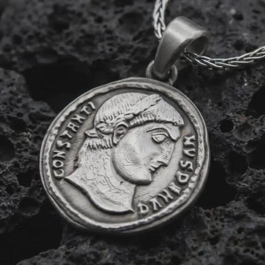 Oxidized Emperor Constans coin pendant necklace resting on black volcanic stone, featuring a Roman imperial profile with Latin inscription in a detailed antique coin-style macro photograph.