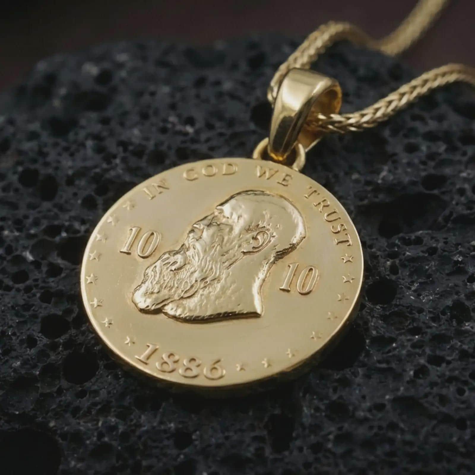 Gold plated heraldic eagle coin pendant necklace photographed on porous black volcanic stone, emphasizing the regal emblem, round medallion silhouette, and symbolic laurel accents in a luxury jewelry composition.