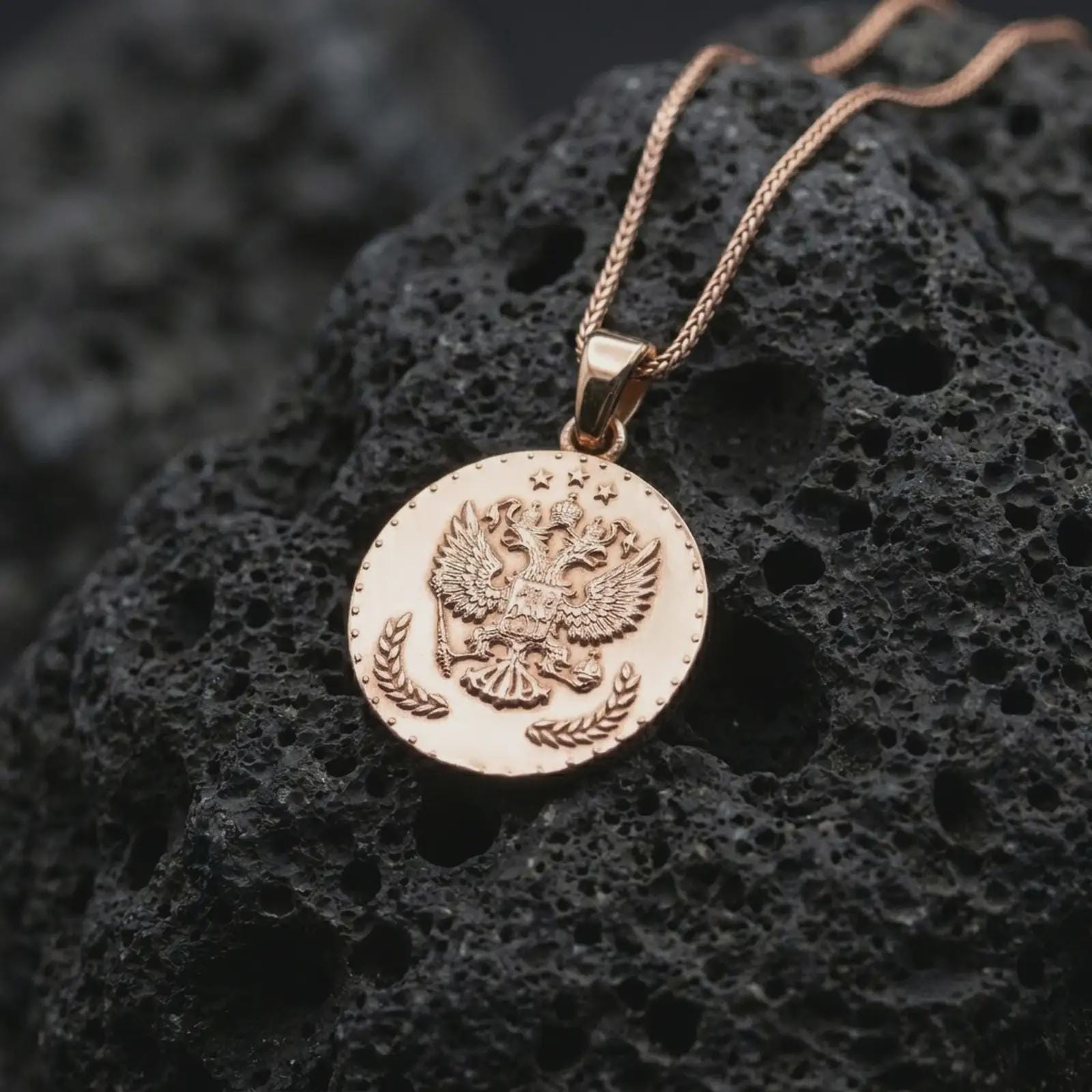 Rose gold plated heraldic eagle coin pendant necklace presented on textured volcanic stone, showcasing the crowned eagle crest and coin-inspired relief in a refined macro studio shot.