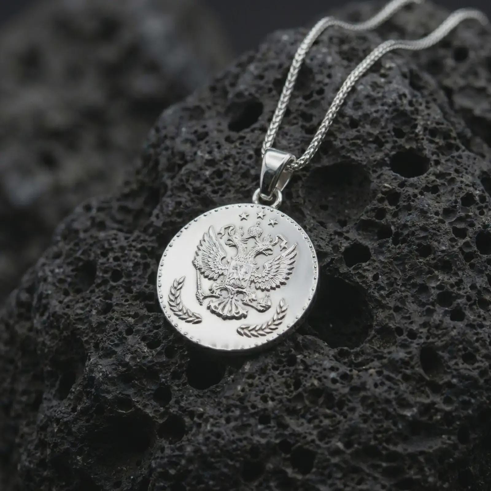 Polished silver heraldic eagle coin pendant necklace displayed on dark volcanic rock, shown in a premium close-up image that highlights the imperial double-headed eagle motif and antique coin-style detailing.