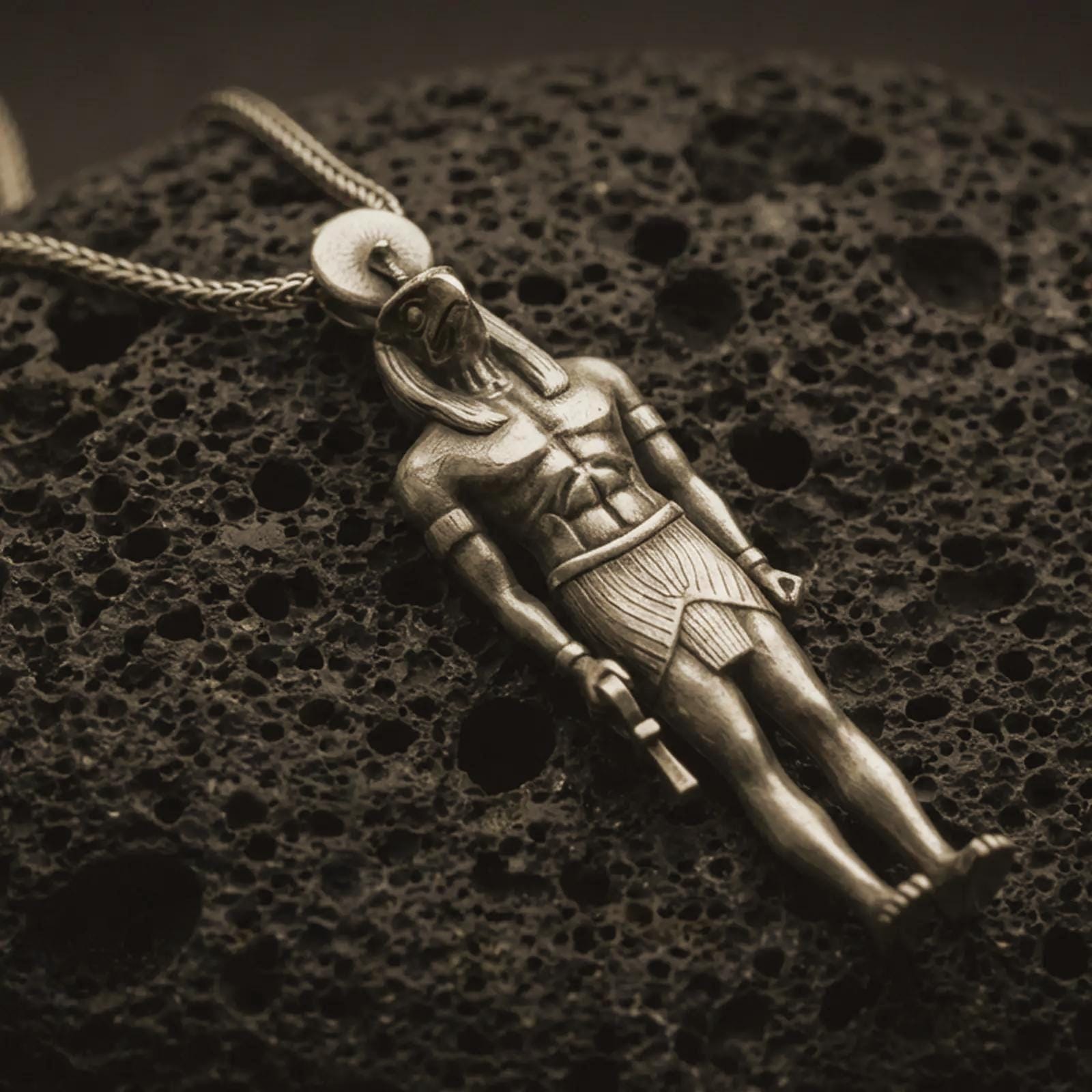 Bronze Ra amulet pendant necklace resting on black volcanic stone, featuring an ancient Egyptian deity figure with a falcon-like head, sun disk, and strong sculpted body in a detailed macro product photograph.