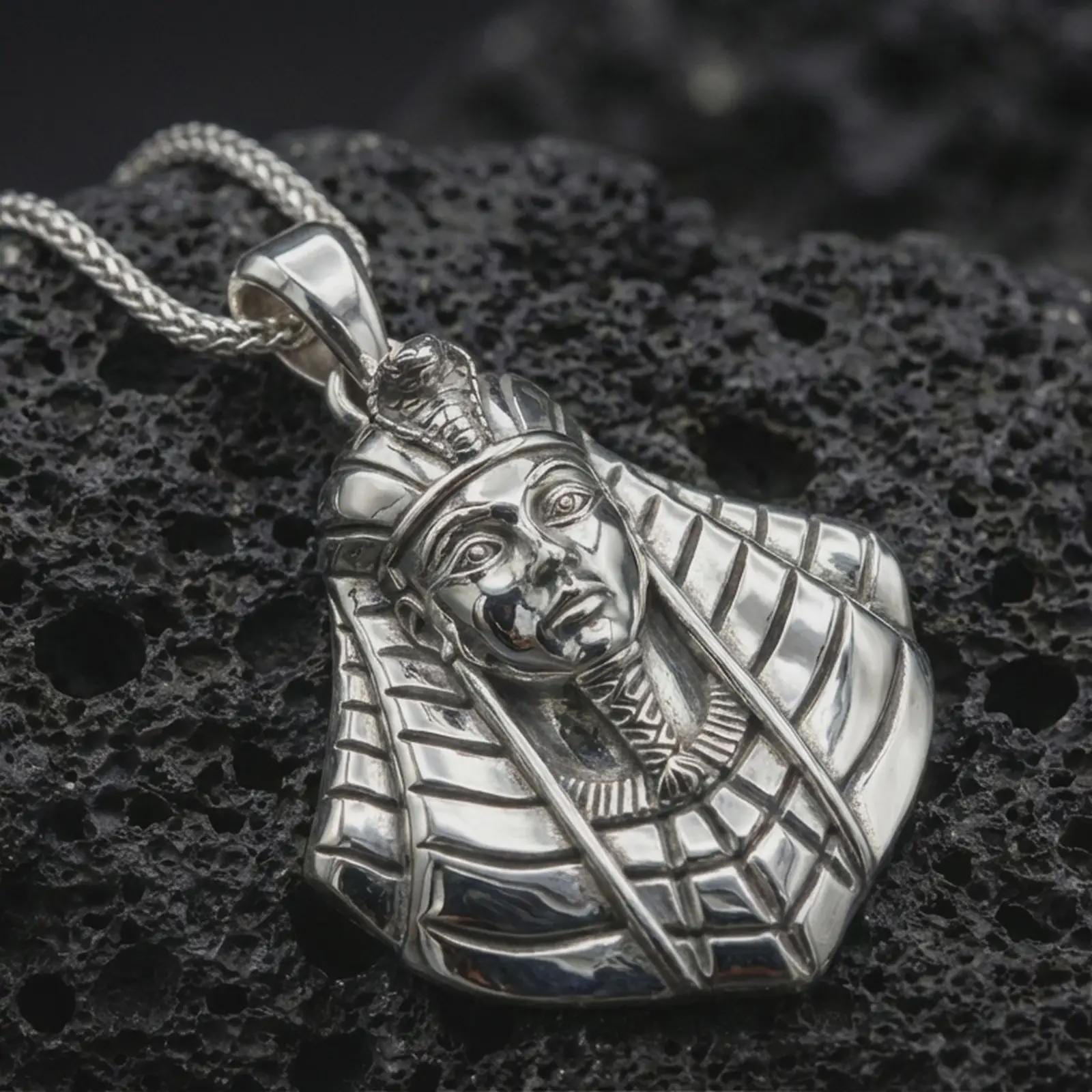 Polished silver Tutankhamun pendant necklace displayed on dark volcanic rock, shown in a premium close-up image that highlights the royal pharaoh face, striped headdress, and ancient Egyptian styling.