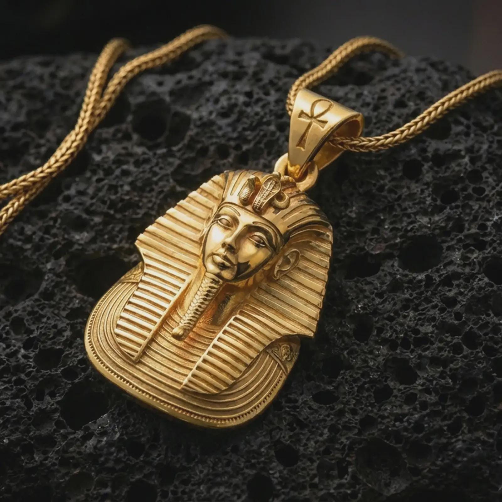 Gold plated Tutankhamun pharaoh pendant necklace photographed on porous black volcanic stone, emphasizing the iconic king portrait, layered headdress lines, and historic Egyptian symbolism in a luxury jewelry composition.