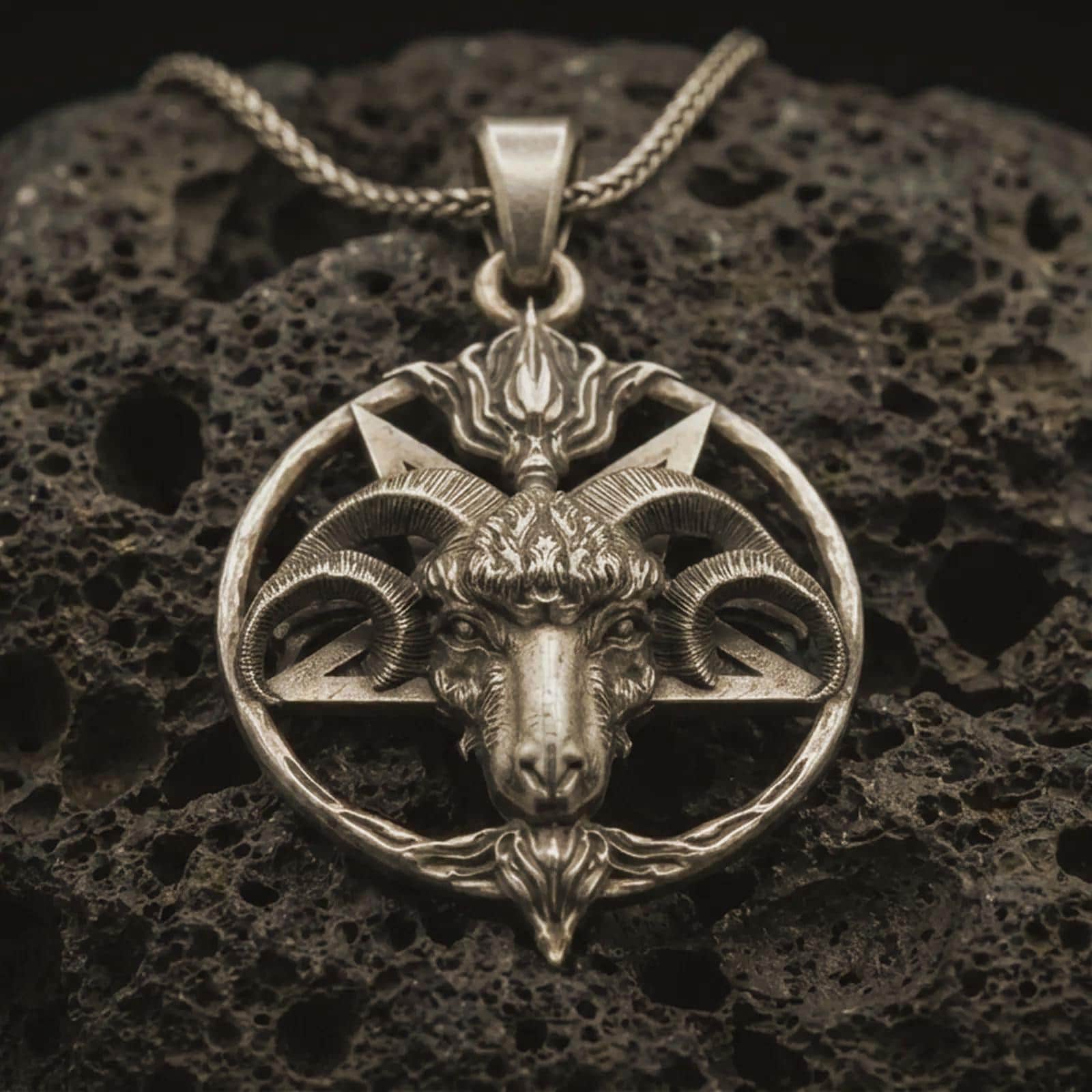 Bronze Baphomet goat head pendant necklace with an oxidized finish resting on textured volcanic rock, captured in a close-up premium studio composition.