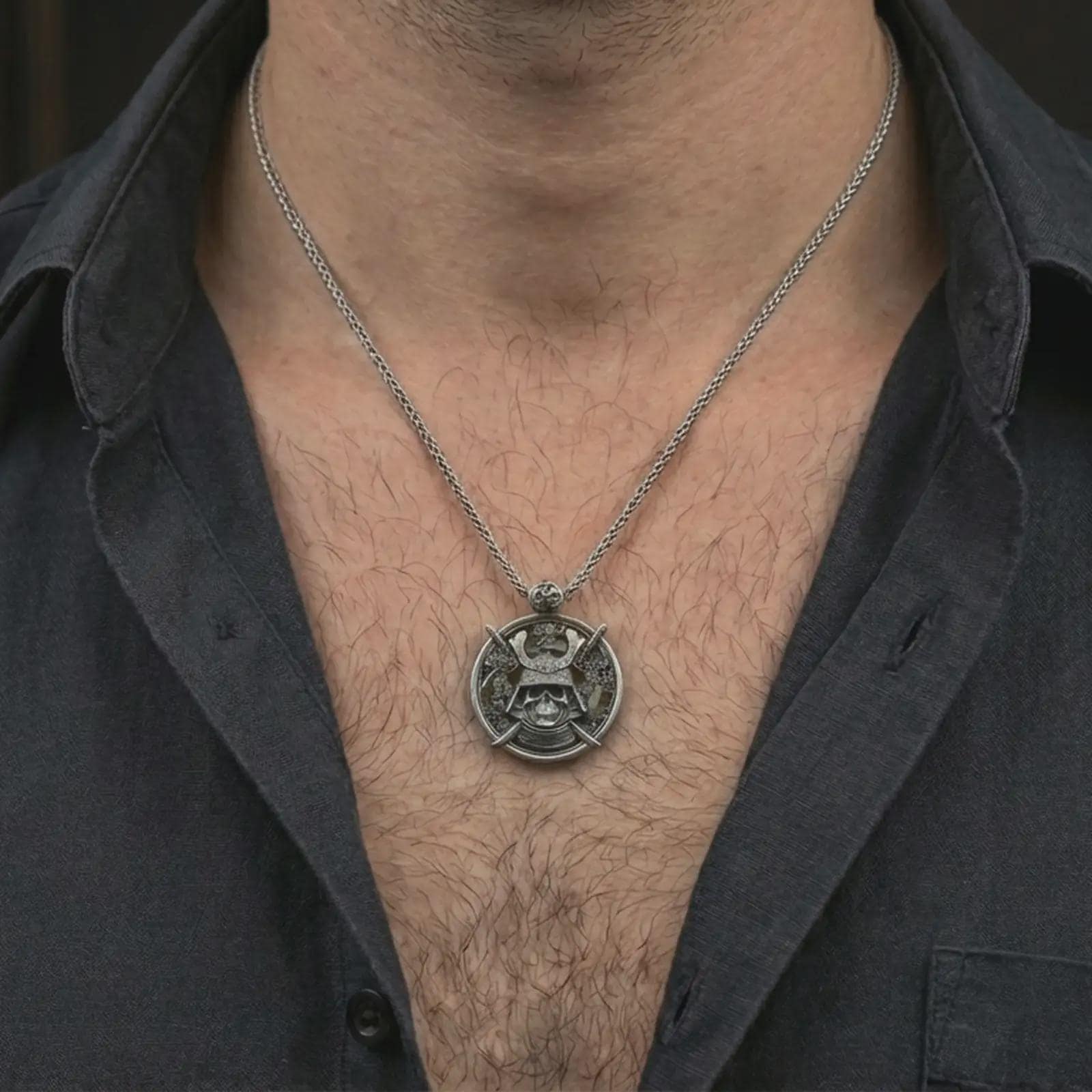 Oxidized samurai skull pendant necklace worn on the neck, styled in a dark lifestyle portrait with an open shirt and masculine presentation.