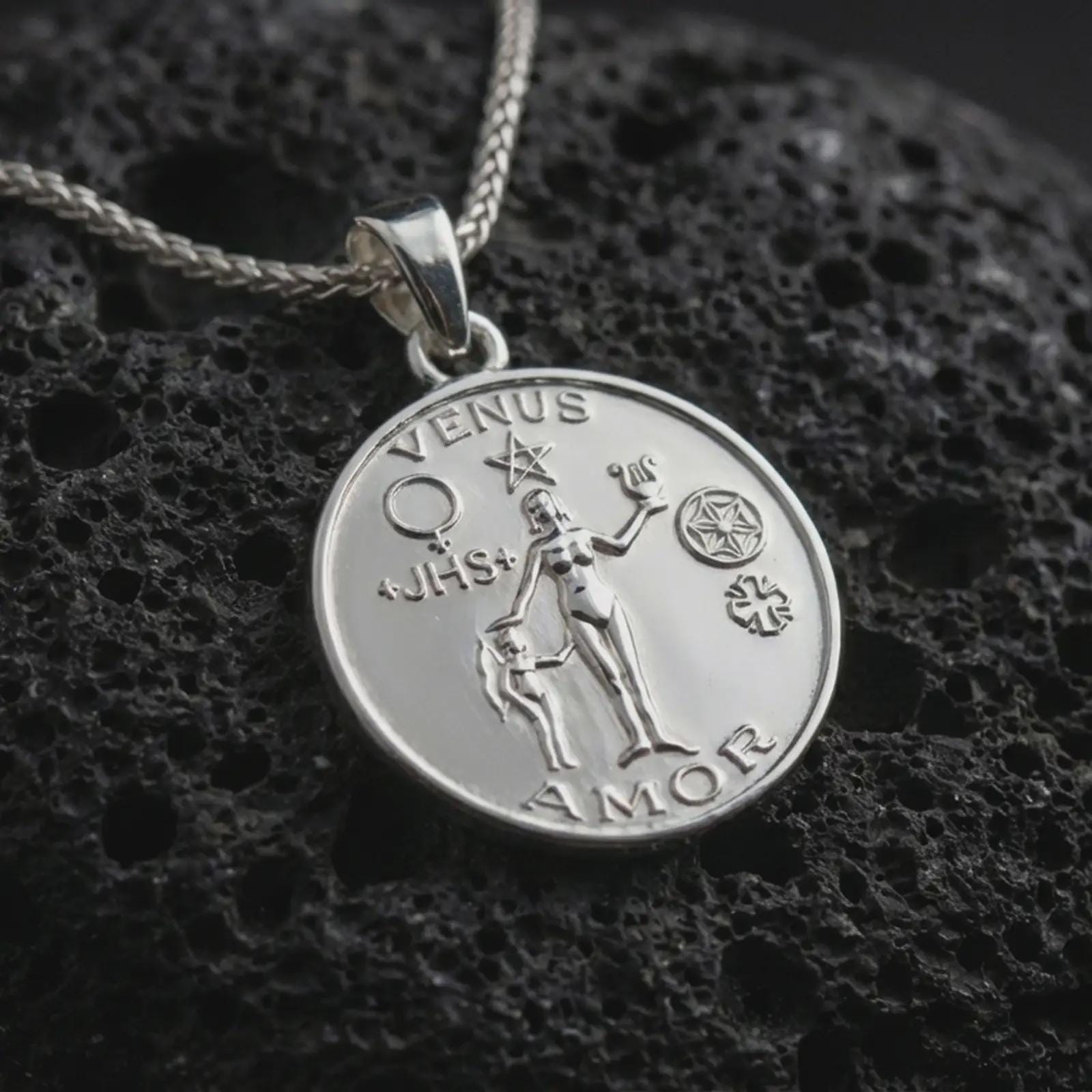 Polished silver Venus Amor pendant necklace displayed on black volcanic rock, shown in a refined macro product shot with goddess figure, symbolic motifs, and engraved medallion detail.