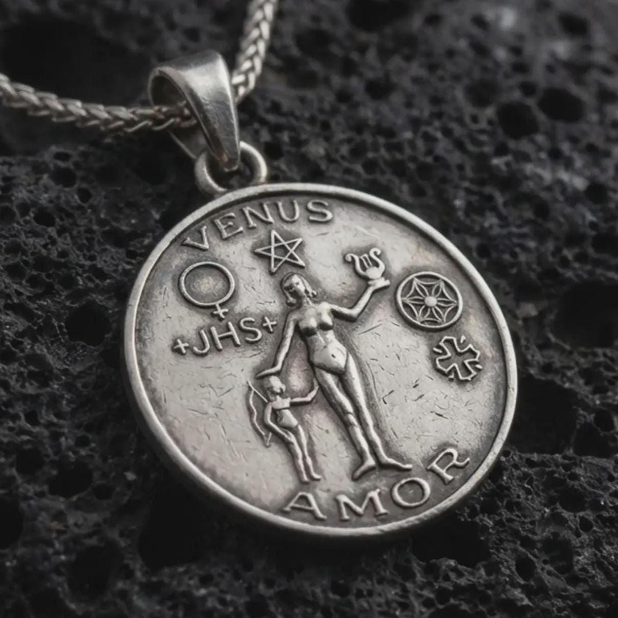 Sterling silver Venus Amor pendant necklace with an oxidized finish resting on textured volcanic rock, captured in a close-up premium studio composition.