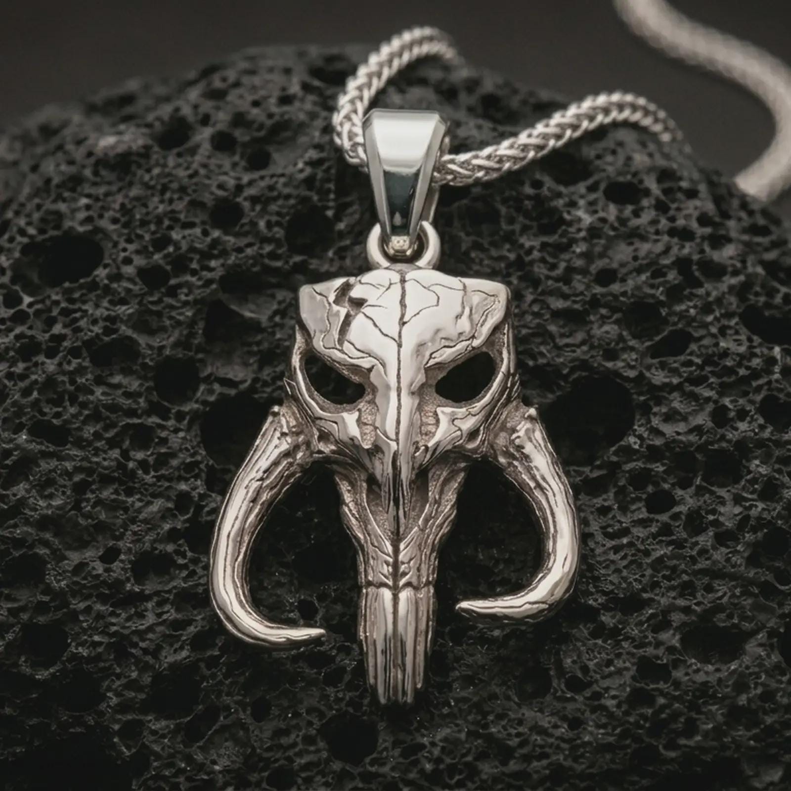 Polished silver Mythosaur skull pendant necklace displayed on black volcanic rock, shown in a refined macro product shot with horned alien skull detail and rugged sci-fi relief.