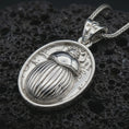 Load image into Gallery viewer, Polished silver Egyptian scarab pendant necklace displayed on black volcanic rock, shown in a refined macro product shot with carved beetle relief, symbolic hieroglyph details, and ankh-inspired bail.
