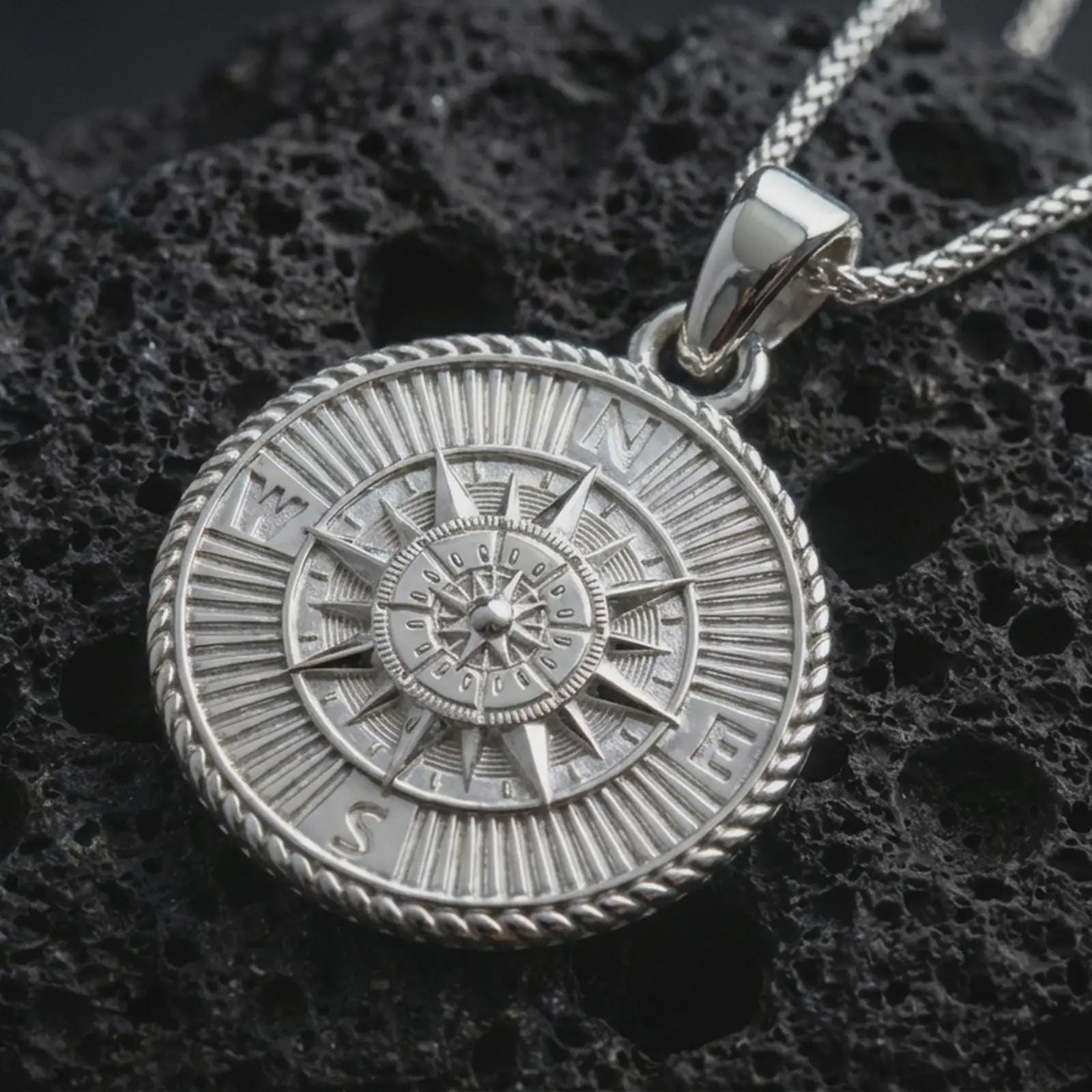 Polished silver steampunk compass pendant necklace displayed on black volcanic rock, shown in a refined macro product shot with detailed directional markers, layered compass rose, and vintage explorer styling.