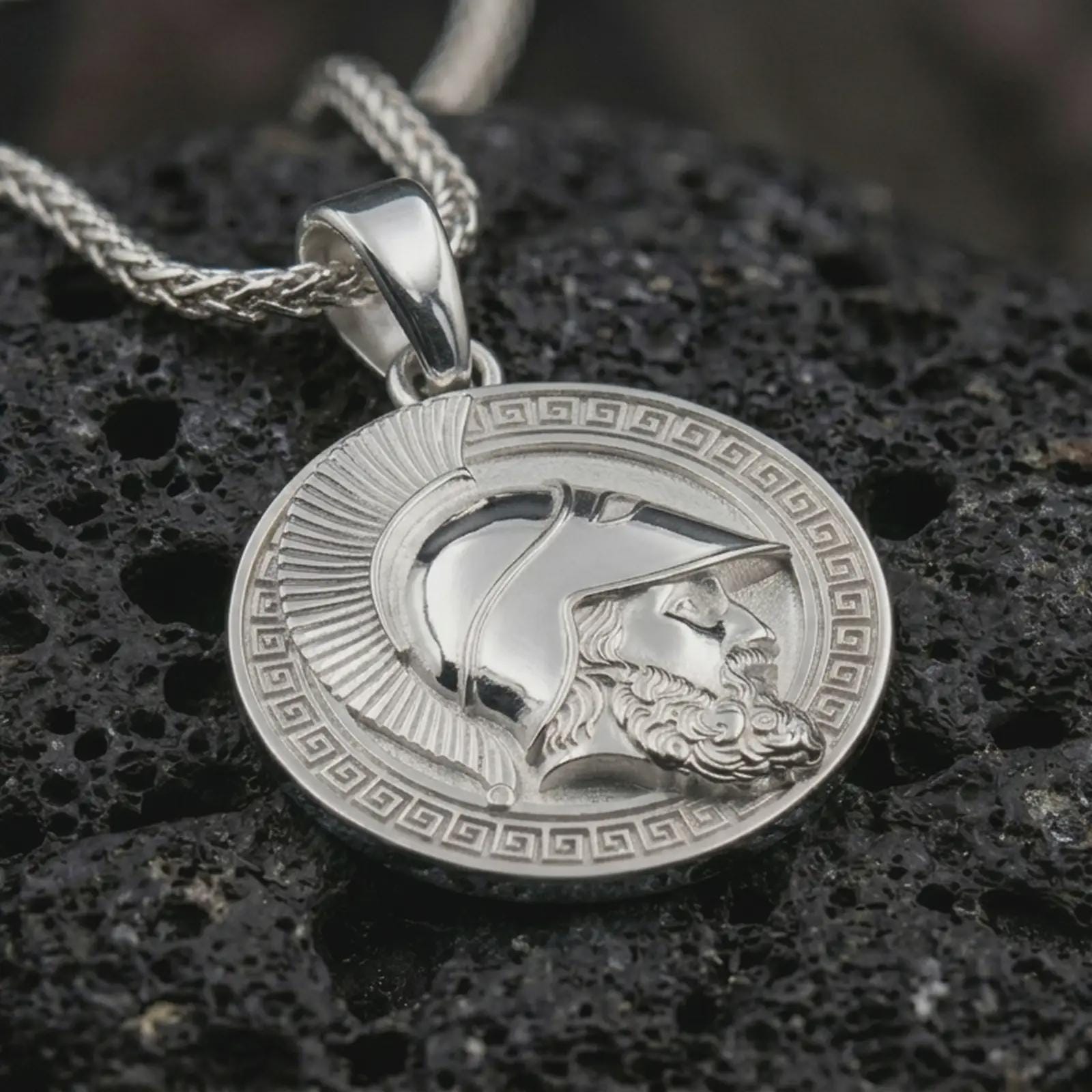 Polished silver Spartan helmet pendant necklace displayed on black volcanic rock, shown in a refined macro product shot with Greek key border, warrior profile, and classical armor detail.
