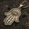 Load image into Gallery viewer, Bronze Hamsa hand pendant necklace with an oxidized finish resting on textured volcanic rock, captured in a close-up premium studio composition.
